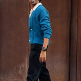 A person wearing a blue cardigan, white shirt, and black pants stands sideways in front of a brown wall, sporting the CASIO G-Shock Watch ANALOG-DIGITAL GA110GB-1A from G-Shock Watches on their wrist while looking slightly away from the camera.