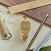 A CASIO Watch A168WG-9VT by G-Shock Watches with a stainless steel bracelet is displayed on wood, surrounded by drawing tools. The vintage digital watch shows the date and time.