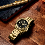 A Seiko Watches SRPK18 Seiko 5 Sports SGP gold automatic watch with a black dial rests on a wooden surface beside a black notebook and gold pen.