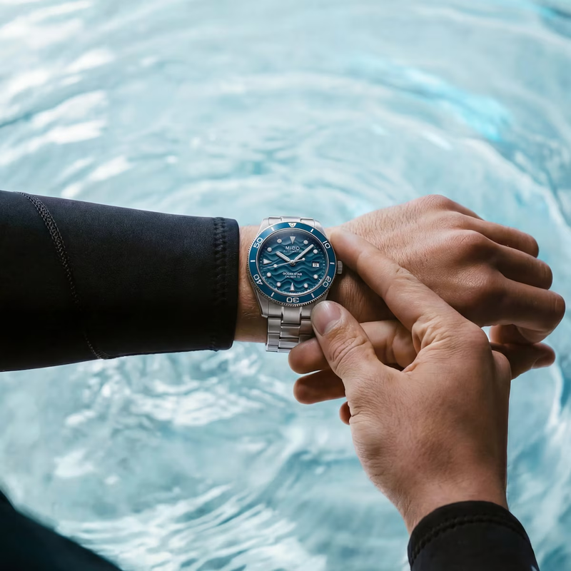 Person wearing a wetsuit adjusting the blue-dial Mido Ocean Star 39 automatic dive watch near swirling blue water