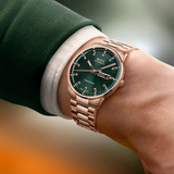 A person in a green suit showcases the Mido Watches Commander Datoday M0214303309100, featuring a rose gold case, green dial, sapphire glass, day-date display, and metal bracelet. The softly blurred background adds elegance.
