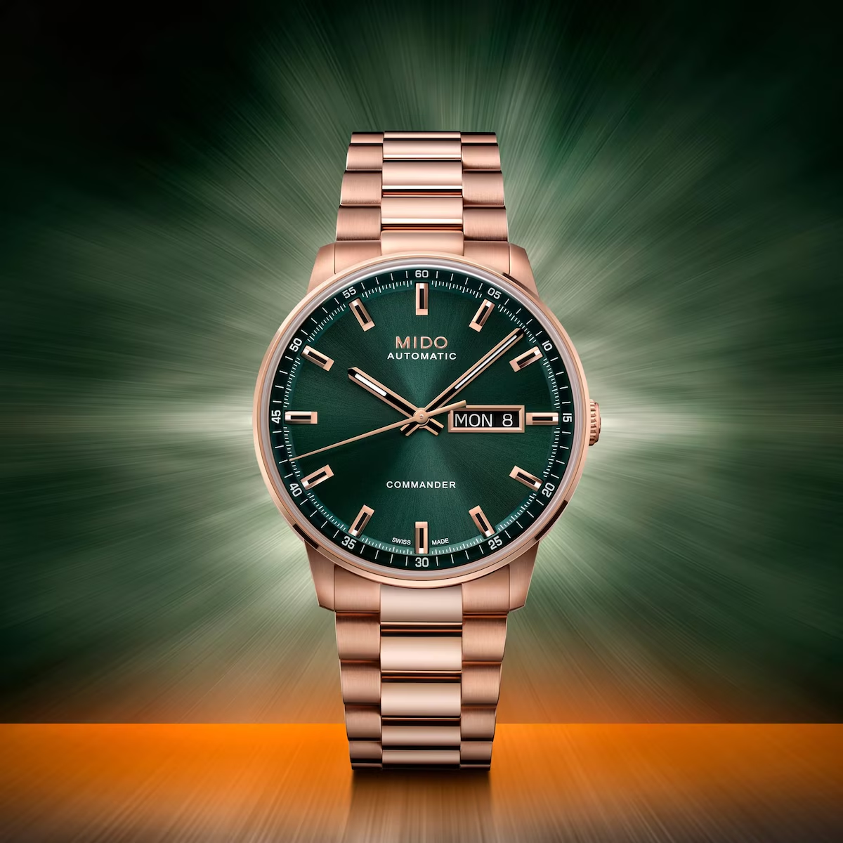 The Mido Watch Commander Datoday M0214303309100 by Mido Watches, featuring a green dial and rose gold PVD finish, is set against a vivid green and orange backdrop, showcasing its day-date display and sophisticated metal bracelet.
