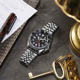 A Seiko Watches SSK001 5 SKX Sports Style wristwatch with a metal bracelet rests on a gray tabletop beside a gold key, brass lamp, pen, and handwritten note on a decorative tray.