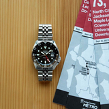 A Seiko Watches SSK001 5 SKX Sports Style, highlighting Japanese craftsmanship, rests on a wooden surface next to several folded King County Metro bus schedules.