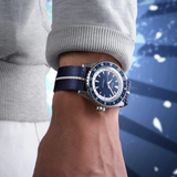 A person wears a Mido Watches OCEAN STAR GMT M0268291804100 with a blue dial and silver bezel, paired with a light gray long-sleeve shirt. The background shows blue tones and blurred lights.