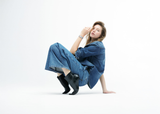 A woman with light brown hair wearing a denim jacket, wide-leg jeans, black heeled boots, and a Frederique Constant Classics Manchette FC-200WR1MC6B watch sits on the floor against a white background, looking at the camera.