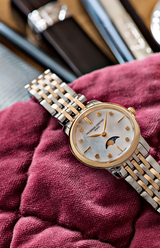 A Frederique Constant Watches Classics Slimline Ladies Moonphase FC-206MPWD1S2B with a gold and silver link band rests on quilted burgundy fabric, featuring a white dial, gold markers, and elegant hands; blurred accessories accent the background.