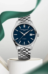 A Frederique Constant Classics Index Automatic FC-303NN5B6B wristwatch with a blue dial and silver markers stands upright on a white pedestal, highlighted by a swirling green ribbon—showcasing Frederique Constant luxury timepieces.
