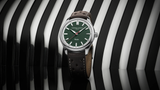 The Frederique Constant Vintage Rally Healey Automatic FC-301HGRS5B26 men’s watch with a racing green dial, silver markers, and brown perforated leather strap stands upright against a black and white wavy striped background.