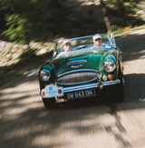 Two people in sunglasses and hats drive a classic dark green convertible with a Frederique Constant Vintage Rally Healey Automatic FC-301HGRS5B26 watch glinting in the sun on a curving, tree-lined road. The photo shows slight motion blur.