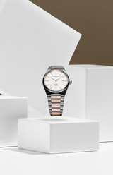 A Frederique Constant Highlife Automatic COSC FC-303V4NH2B watch with a silver and rose gold band sits on a white geometric pedestal, its globe-pattern dial and gold hour markers set against minimalist white blocks and a brown wall.
