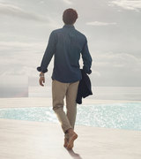 A barefoot man in beige pants and a blue shirt walks toward a pool with the ocean behind him, holding a jacket and wearing a Frederique Constant Highlife Automatic COSC FC-303V4NH2B watch with a globe-pattern dial.