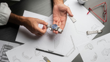 A person holds a Frederique Constant Highlife Automatic COSC FC-303V4NH2B with a blue globe-pattern dial above watch design sketches on a desk, surrounded by straps, a black pen, red glasses, and a white coffee cup.