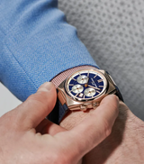 A person in a light blue textured suit adjusts a Frederique Constant Highlife Chronograph Automatic FC-391NS4NH4 watch with a blue face, three subdials, dark strap, and visible crown on their left wrist.