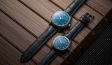 Two Frederique Constant Classics Runabout Automatic FC-303RMLN5B6 Limited Edition watches, featuring blue dials, silver cases, and black leather straps with blue stitching, are arranged diagonally on a wood-textured surface.