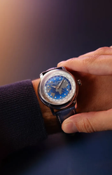 A hand in a dark blue sweater showcases the Frederique Constant Manufacture Classic Worldtimer FC-719BLW3H6 watch, featuring a blue world map dial and dark leather strap, by Frederique Constant, against a blurred dark background.