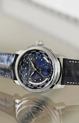 The Frederique Constant Manufacture Classic Worldtimer FC-718NWM4H6 features a dark blue and silver world time dial with city names, multiple time zones, and a black alligator leather strap, displayed on a reflective surface.