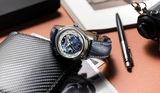 A Frederique Constant Manufacture Classic Worldtimer FC-718NWM4H6, a Swiss automatic watch with a blue leather strap, sits on a desk next to a textured wallet, pen, camera, and a partially visible black and white photo book.