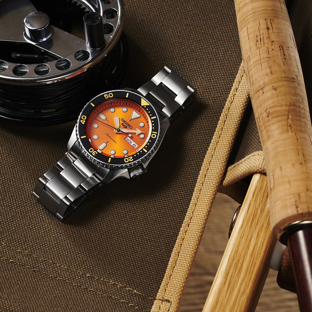 A Seiko Watches 5 Sports SKX series SRPD59 automatic watch with an orange dial is displayed on olive-green fabric next to fishing gear, including a fishing reel and a wooden-handled rod.