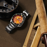 A Seiko Watches 5 Sports SKX series SRPD59 automatic watch with an orange dial is displayed on olive-green fabric next to fishing gear, including a fishing reel and a wooden-handled rod.