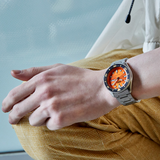 A person in yellow pants wears a Seiko Watches 5 Sports SKX series SRPD59 with an orange dial and black bezel, resting their hand on their knee against a softly blurred background.