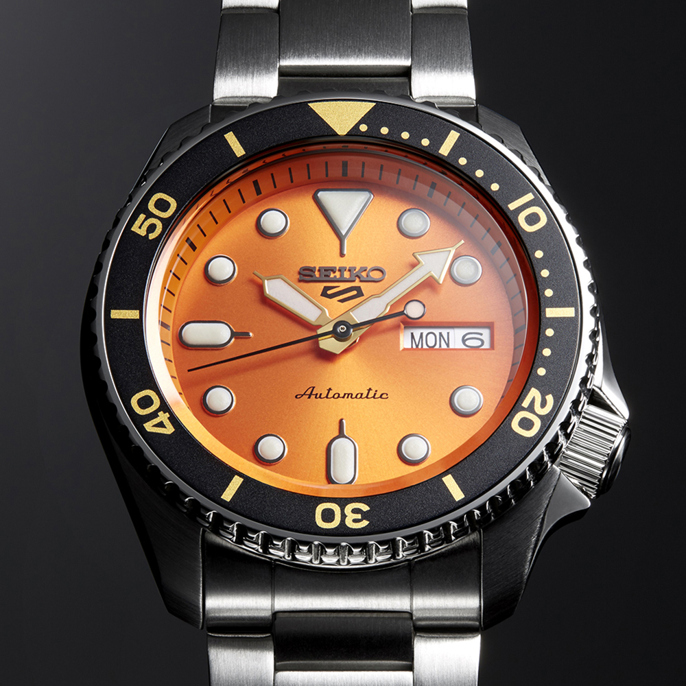 The Seiko Watches 5 Sports SKX series SRPD59 features an automatic movement, stainless steel band, orange dial, large luminous markers and hands, black bezel with yellow markings, and a day-date window displaying MON 6.