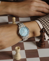 A wrist wearing the Bulova Watches Marine Star 96P248 Ladies (UPC: 042429597433) with diamond hour markers and a light blue face rests on a chessboard amid pieces, complemented by a brown and white houndstooth sleeve.