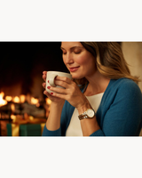 A woman with wavy blonde hair, in a blue sweater, smiles gently holding a white mug near her face while wearing the Bulova Watches Marine Star 98P227 Ladies watch, sitting in front of a cozy fireplace.
