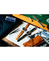 Two wristwatches, including the Bulova Watches Chronograph A 98B427 Men with perforated leather straps, are displayed on papers beside a Bulova card, tools, a metal band, and a brown leather pouch on a wooden surface.