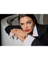 A woman with slicked-back dark hair and bold red lipstick, wearing a black blazer with white cuffs, gold jewelry, and the Bulova Watch Sutton 97L177 by Bulova Watches, leans on a white staircase rail and looks seriously at the camera.