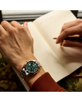 Wearing a Bulova Watches Surveyor 96B429 men’s watch with a green dial and stainless steel band, someone writes in an open notebook with a pen, showing just their hands and the brown sleeve of their shirt.