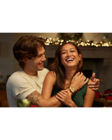 A cheerful couple embraces in a festive, warmly lit room. The man, in a white shirt and wearing the Bulova Watches Surveyor 97A182 Men by Bulova Watches, holds the woman from behind as she laughs joyfully.