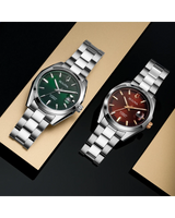 Two Bulova Surveyor Men's Watches (model 98B422) with stainless steel bands are shown on gold and black surfaces—one with a green automatic dial, the other with a brown face, gold crown, and silver hour markers.