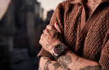 A person in a brown textured short-sleeve shirt and a Bulova Watches Bulova 98B451 watch crosses their tattooed arms, with a blurred cityscape in the background.