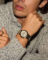 A person in a textured gray knit sweater rests their chin on their hand, showcasing a Bulova Prestige 98B450 watch by Bulova Watches in gold and silver stainless steel, along with a gold ring featuring a decorative design.