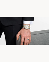 A person in a black suit with a white shirt cuff wears a Bulova Prestige 98B450 watch (UPC 042429603363) from Bulova Watches, gold and silver stainless steel, with a gray ring, resting their hand on a light gray upholstered chair.