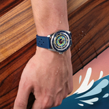 A wrist sports the Mido Watches SPECIAL EDITION OCEAN STAR DECOMPRESSION WORLDTIMER M026.829.17.041.00 with a blue strap and colorful dial, water droplets on the hand, near wood and an illustrated water splash in the bottom right.