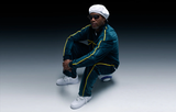A person in a white beret, Dita Sunglasses DLS404-56-02-A by Dita Sunglasses, a teal tracksuit with yellow stripes, and white sneakers squats on the ground against a gradient dark background.
