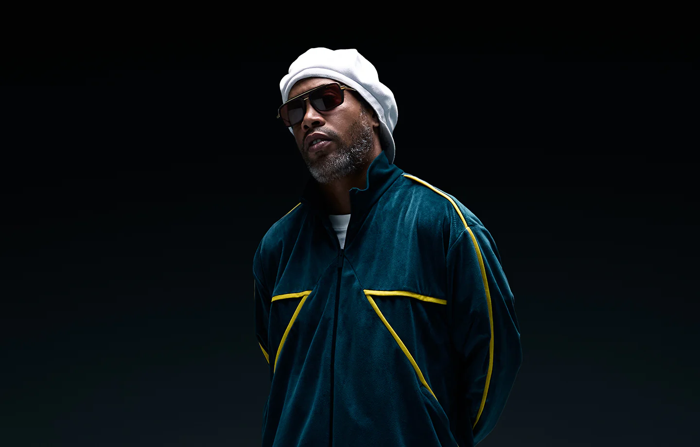 A man in a dark green jacket with yellow accents, a white beret, and Dita Sunglasses DLS404-56-02-A LSA-404 stands against a high-contrast dark background, looking confidently at the camera.
