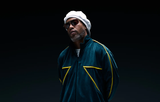 A man in a dark green jacket with yellow accents, a white beret, and Dita Sunglasses DLS404-56-02-A LSA-404 stands against a high-contrast dark background, looking confidently at the camera.