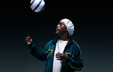 A man in Dita Sunglasses DUBSIGNAL DTS465-A-03 Unisex, a white beanie, and green tracksuit looks up at a blue and white soccer ball about to head it, set against a dark background. The stylish Dita eyewear adds unisex appeal.