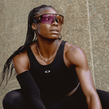 A woman in a black Oakley top, braided hair, and silver chain necklace crouches outdoors before a textured wall, wearing Oakley Meta Vanguard Sunglasses 0OW8001 800109 by Oakley Meta Sunglasses as she looks up and to the side.