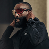 A man with a full beard wears Oakley Meta Sunglasses 0OW8001 800109, a black hooded jacket, and a smartwatch, adjusting his hood as he looks ahead with focus.