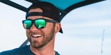 A man wearing a black cap and Costa Del Mar Sunglasses 900818 Spearo Matte Black smiles while on a boat. The reflection of water and part of the boat can be seen in the polarized lenses of his Costa Del Mar Sunglasses. The background is bright and slightly blurred.