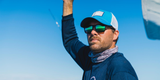A man wearing a blue cap, Costa Del Mar Sunglasses 900818 Spearo Matte Black, and a neck gaiter raises his arm against a bright blue sky.