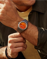 A person adjusts a Citizen Watches AW1840-50X Brycen with an orange dial on their left wrist. They are dressed in a khaki jacket layered over a beige shirt, and wear a black bracelet on their right wrist. The silver watch, crafted from Super Titanium and powered by Citizen’s Eco-Drive technology, stands out as both stylish and innovative.