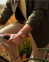 A person dressed in tan pants, a beige shirt, and an olive jacket holds onto a bicycle handlebar. On their wrist is the Citizen Watches AW1840-50X Brycen by Citizen Watches (UPC 013205165979), featuring Eco-Drive technology, a silver case, and an orange face. Sunlight illuminates the greenery in the background.