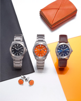 Three Citizen Watches, including the bold Citizen Watches AW1840-50X Brycen, are showcased with striking black, orange, and blue dials on a color-blocked backdrop, accompanied by orange cufflinks and an orange wallet. These timepieces feature innovative Eco-Drive technology and are expertly crafted from Super Titanium® with sleek metal bands—while one model is enhanced by a classic brown leather strap—uniting modern style with enduring durability.