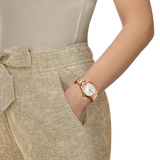 A person wearing beige linen pants and a light top has their hand in their pocket, showcasing a Tissot Watches PRX 34mm T1370101135100—a refined Swiss Made women’s watch with a white face and rose gold stainless steel—on their left wrist.