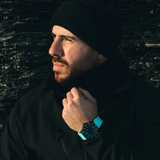 A man wearing a black beanie and jacket looks to the side, holding his collar with one hand. He is outdoors and sporting the Luminox Watches Pacific Diver Limited Edition 44 mm XS.3124.N.SET in teal on his wrist. The background is dark and textured.